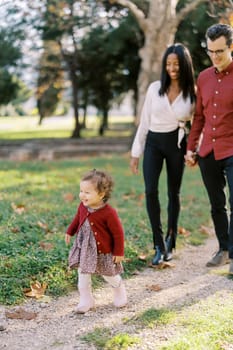 Dad and mom walk holding hands behind a little smiling girl along a path in the park. High quality photo