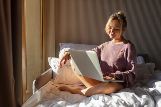 Happy casual beautiful woman working on a laptop sitting on the bed in the house