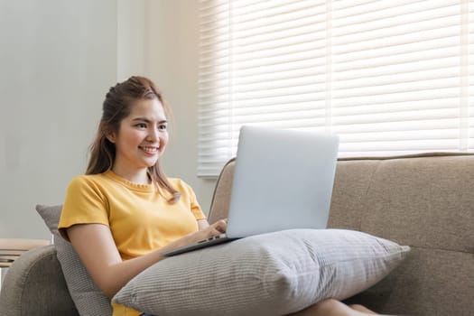 Beautiful Asian woman wearing casual clothes On the sofa using a laptop computer, entertaining with social media, relaxing, smiling and laughing..