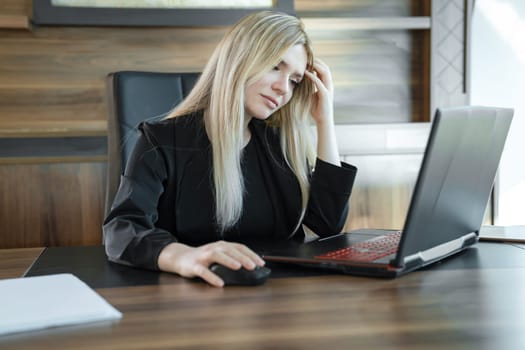 Focused girl blonde in black suit is sitting front of laptop in office, concept of online education and work.