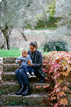 Mom with a little girl on her knees sits on stone steps in the park and shows her a red leaf in her hand. High quality photo