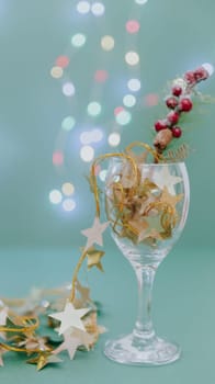 One wine glass with a garland of golden stars and a fir branch in it stands on a pastel green background with blurred bokeh from the burning garland, close-up side view.