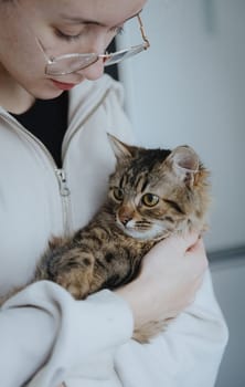 Portrait of one beautiful Caucasian teenage girl in glasses and a light jacket cutely holding a small purebred kitten in her arms, standing in the room on a winter day, flat lay close-up.