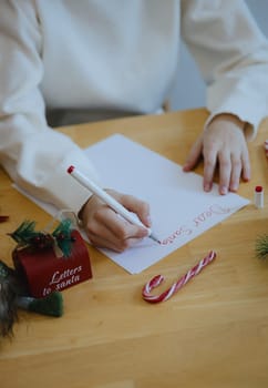 One unrecognizable Caucasian teenage girl writes a letter to dear Santa Claus with a red felt-tip pen, sitting at a wooden table with Christmas decorations, in the dining room on a winter day, side view from above, close-up.