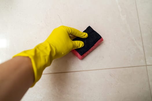 A hand in a yellow glove holds a sponge and wipes the tiles on the wall in the bathroom, a man does cleaning with detergent at home.