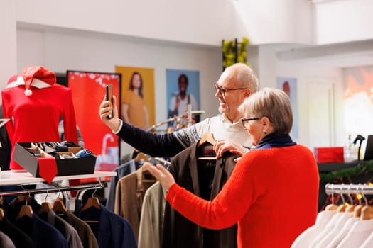 Elderly couple taking photos in store, comparing jackets on hangers with products online on website. Senior people shopping for formal clothes during christmas season, looking for festive clothing.