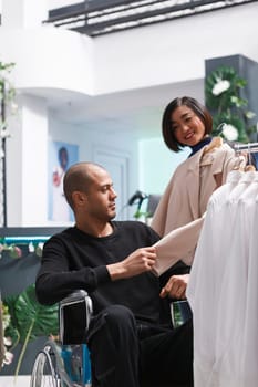 Arab man in wheelchair receiving guidance from asian woman seller while shopping for formal apparel. Boutique customer with disability examining beige jacket while consultant holding hanger