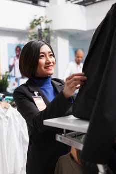 Clothing store smiling asian employee fastening jacket buttons while wearing mannequin in formal apparel. Shopping mall boutique woman worker organizing womenswear for sale