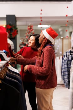 Retail worker showing items to client for shopping assistance during christmas season sales, recommending right clothes size for holiday event preparations. Diverse women discussing fashion products.