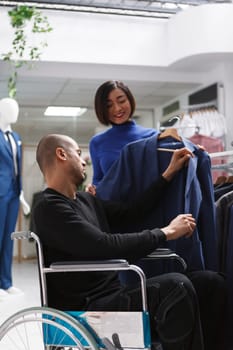 Clothing store asian woman worker providing support while arab man with physical disability checking jacket on hanger. Boutique seller assisting customer in wheelchair to explore apparel