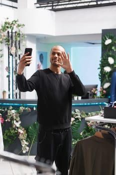 Shopping mall boutique arab man client waving hi while making selfie on smartphone. Young smiling blogger greeting social media followers while live broadcasting in clothing store