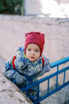 Little girl in overalls stands leaning forward on the fence near the house. High quality photo