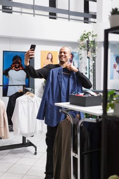 Smiling arab fashion influencer using smartphone front camera while showing outfit on hanger and promoting apparel brand in boutique. Clothing store client taking selfie on mobile phone