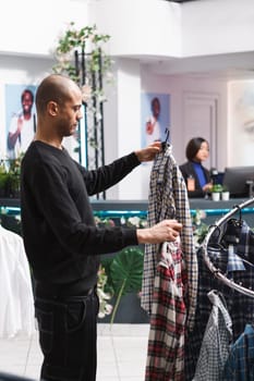 Young arab man holding two plaid shirts on hangers while shopping for casual outfit in shopping mall fashion department. Clothing store client examining garment style and size