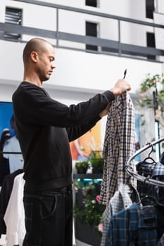 Young arab man in clothing store examining plaid shirt on hanger, checking fit and style. Shopping mall boutique customer browsing apparel rack and checking casual outfit