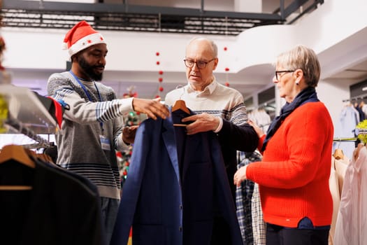 Man employee helping clients to choose jackets and blazers, making preparations for upcoming christmas event. Senior couple shopping for clothes in retail store, buying merchandise on sale.