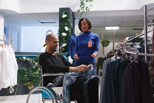 Inclusive clothing store asian woman assistant providing guidance to customer in wheelchair. Arab man in wheelchair buying apparel and getting fashion advice from boutique seller