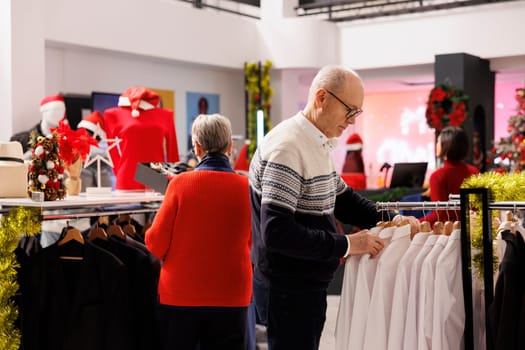 Senior people searching for jackets in shopping mall, looking at clothing items on hangers during winter sales season. Couple checking new clothes in retail store, buying gifts for family.