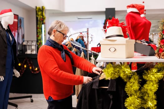 Old customer browsing for formal attire to wear at christmas dinner, stylish blazers on hangers in xmas decorated shopping center. Cheerful woman enjoying shopping session during holidays.