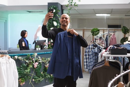 Smiling arab man blogger promoting fashion brand while recording video on smartphone in clothing store. Cheerful influencer showing jacket to social media channel followers