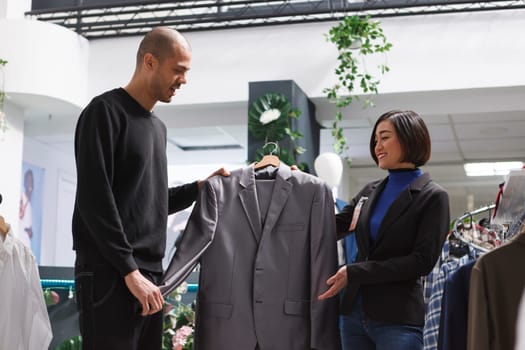 Clothing store asian woman assistant offering advice to an arab man customer, showing formal jacket. Shopping center fashion department consultant and client holding trendy apparel on hanger