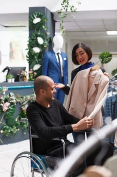 Clothing store friendly assistant discussing outfit choice with client in wheelchair and helping with stylish jacket purchase. Boutique asian woman seller assisting arab man customer with disability