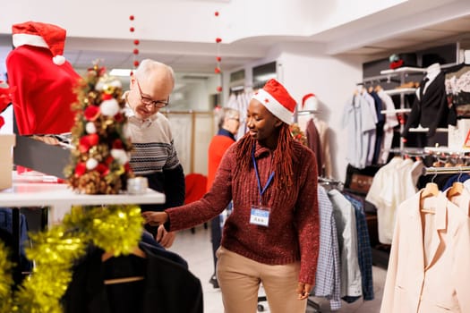 Retail worker helping customer with perfect clothing measurement, recommending fashion item from hangers. Woman store assistant choosing clothes models for senior client, christmas shopping.