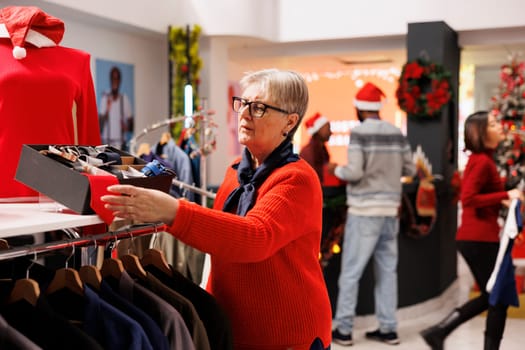 Client checking accessories box in clothing store, searching for ties and belts to give presents on christmas eve celebration. Elderly woman browsing through clothes on hangers, winter promotions.