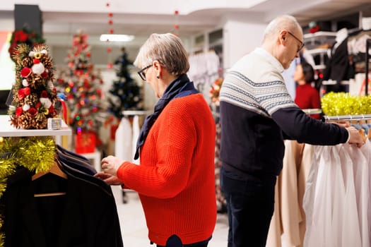 Elderly couple searching for blazers in clothing store with festive decorations, buying items as presents for family on christmas eve. Senior man and woman browsing through merchandise on discount.