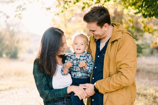 Smiling dad and mom looking at a little girl in her arms while standing in the forest. High quality photo