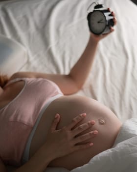 A pregnant woman lies in bed and holds an alarm clock. Close up of the belly