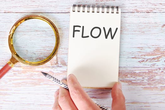 FLOW written on a blank sheet of a notebook which is touched by a man's hand with a pencil and a magnifying glass is lying next to it