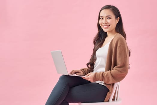 Happy young woman working online, sitting on chair and using laptop against pink wall