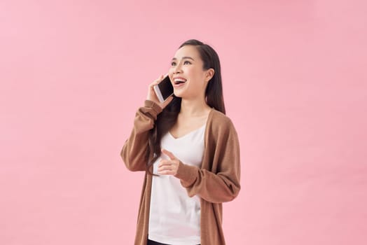 Positive young asian female smiling and talking smartphone while sitting on chair on pink background
