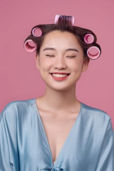 Young pretty asian girl having hair curlers on her head isolated on pink background