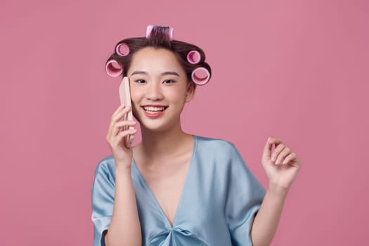 Laughing young woman in hair curlers talking by phone on light background