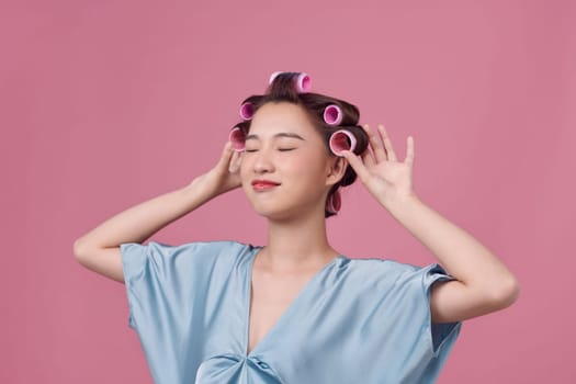 young beautiful girl having hair curlers on her head isolated on pink background