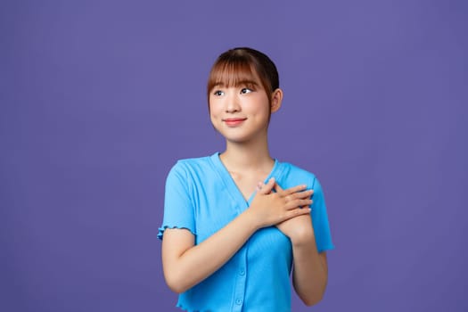 Cheerful woman smiling and holding hands on chest isolated over purple wall