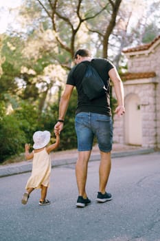 Dad leads a little girl by the hand along the road in the park. Back view. High quality photo