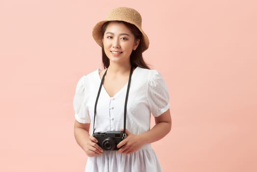 A smiling young woman in summer hat standing with photo camera isolated over pink background