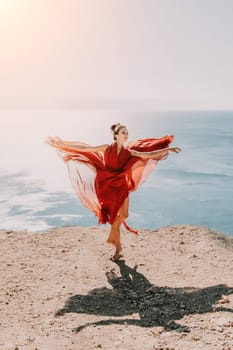 Side view a Young beautiful sensual woman in a red long dress posing on a rock high above the sea during sunrise. Girl on the nature on blue sky background. Fashion photo.