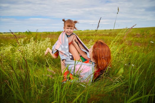 Happy female family with mother and daughter on green and yellow meadow full of grass and flower. Woman with red hair and blonde girl having fun, joy and hug in sunny summer day. Concept family love