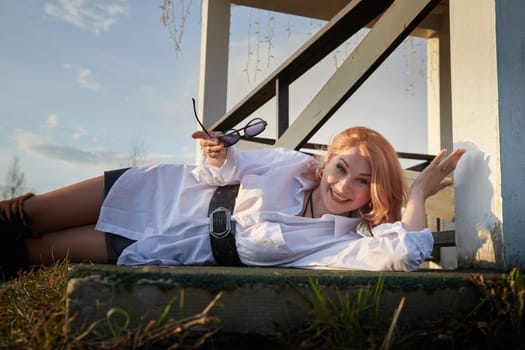 Beautiful girl with red hair in white shirt in open wooden pavillion in village or small town. Young slender woman and sky with clouds on background on autumn, spring or summer day