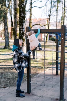 Smiling mother helping little girl climb chain ladder at playground. High quality photo