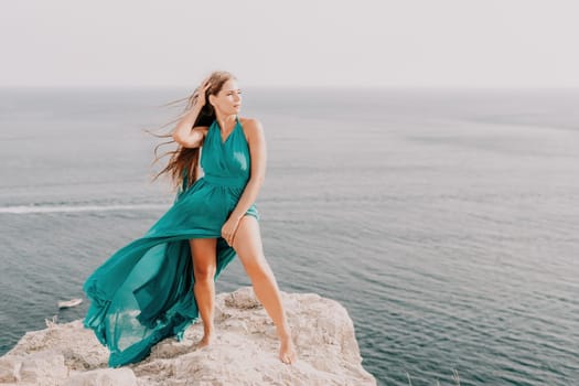 Side view a Young beautiful sensual woman in a mint long dress posing on a volcanic rock high above the sea during sunset. Girl on the nature on overcast sky background. Fashion photo