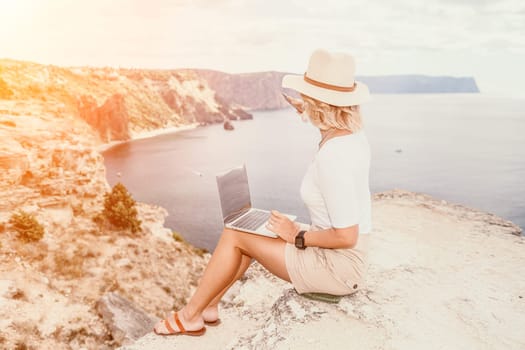 Digital nomad, Business woman working on laptop by the sea. Pretty lady typing on computer by the sea at sunset, makes a business transaction online from a distance. Freelance remote work on vacation