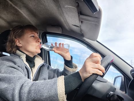 Portrait of female driver in solo journey. Adult mature woman holding steering wheel and bottle of water. Drinking while driving for to stay awake. Lady girl drinks alcohol vodka or gin while driving