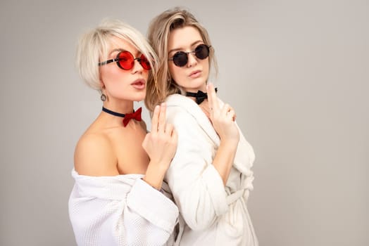 two close friends in white bathrobes and sunglasses stand close to each other and hold their fingers in the shape of a pistol