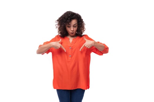 portrait of a bright young caucasian brunette woman with curly hair dressed in an orange shirt pointing her hand in surprise.