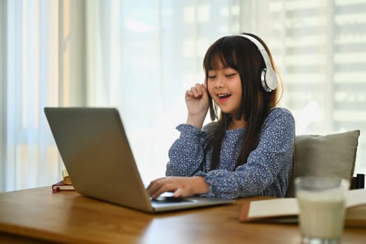 Smiling Asian little cute girl in headphone watching online lesson on laptop at home